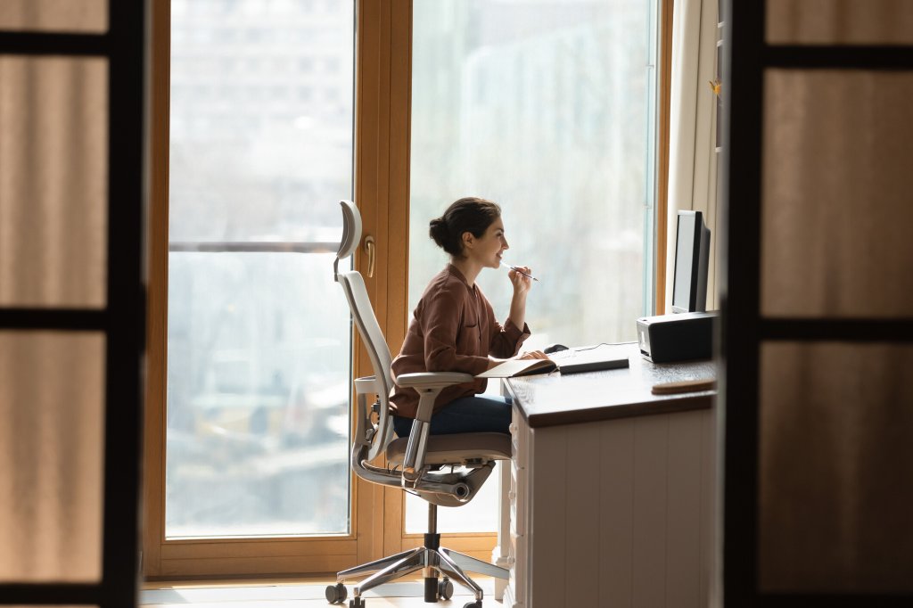 Frau mit braunen Haaren und brauner Blue sitzt auf einem ergonomischen Bürostuhl vor einem Schreibtisch und guckt auf den PC-Bildschirm.