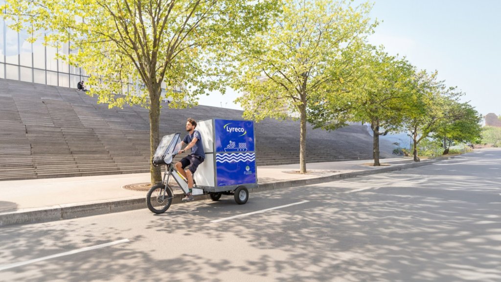 Eine Person fährt mit einem Elektro-Lieferfahrrad von Lyreco auf einer baumgesäumten Straße, symbolisiert nachhaltige urbane Logistik.