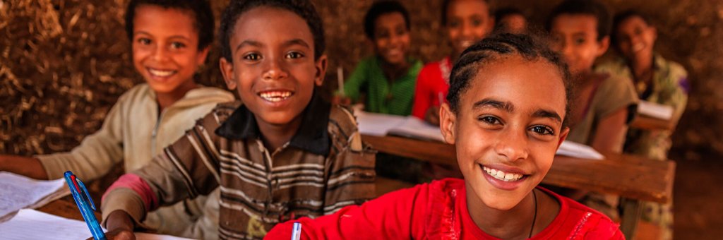 Kinder in einem kambodschanischen Klassenraum, sitzend vor Papier mit Schulaufgaben. Mädchen im Fokus lächelt in die Kamera.