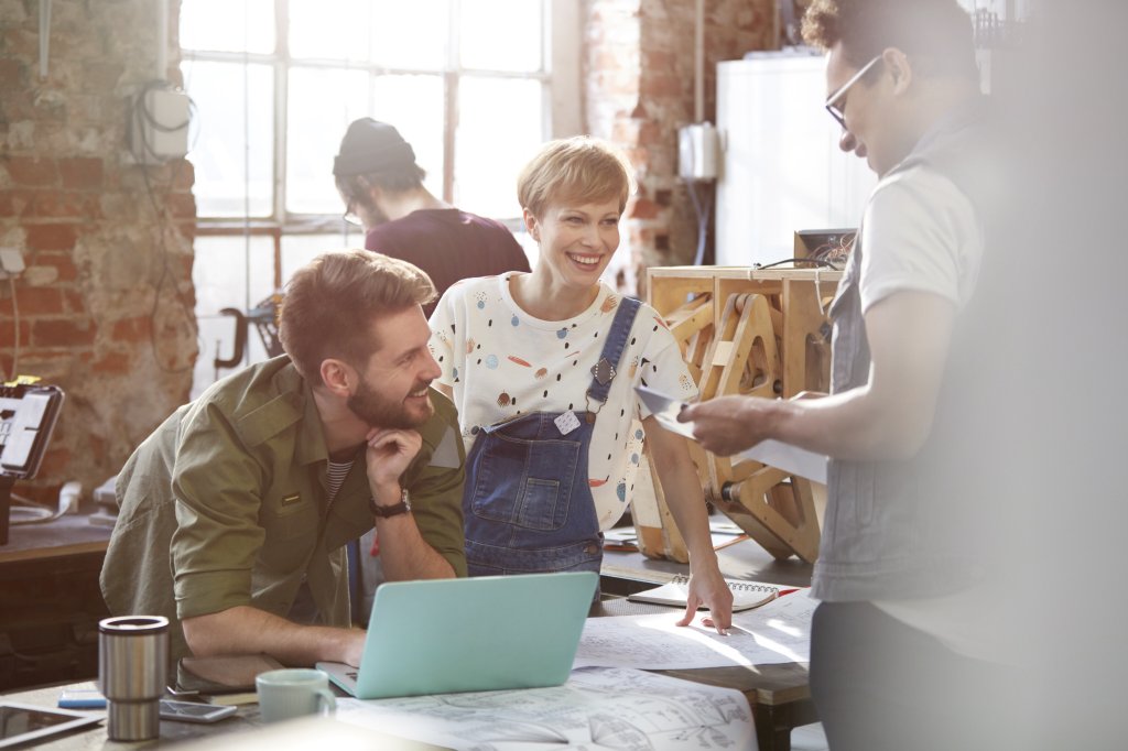 Personen in einem industriellen Büro, an einem Tisch, auf dem sich Zeichnungen und Pläne befinden. Die Personen gucken sich an und lächeln.