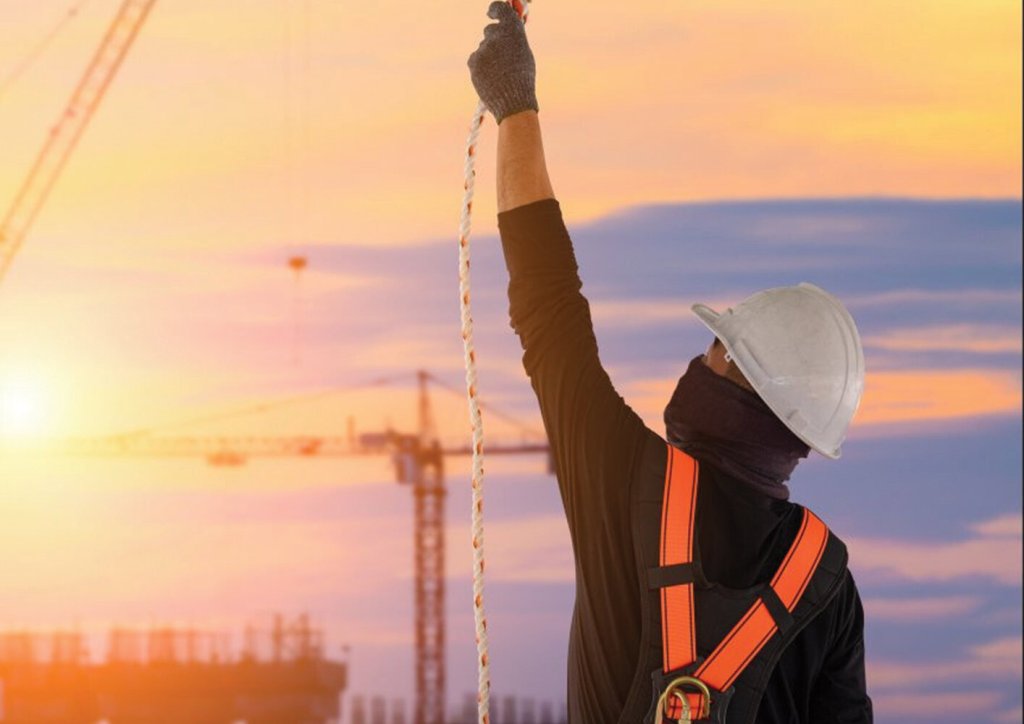 Mann in schwarzer Kleidung trägt weißen Sicherheitshelm und orangenen Fallschutzgurt sowie Sicherheitshandschuhe und greift nach einem Seil. Im Hintergrund befindet sich ein Kran im Sonnenuntergang.