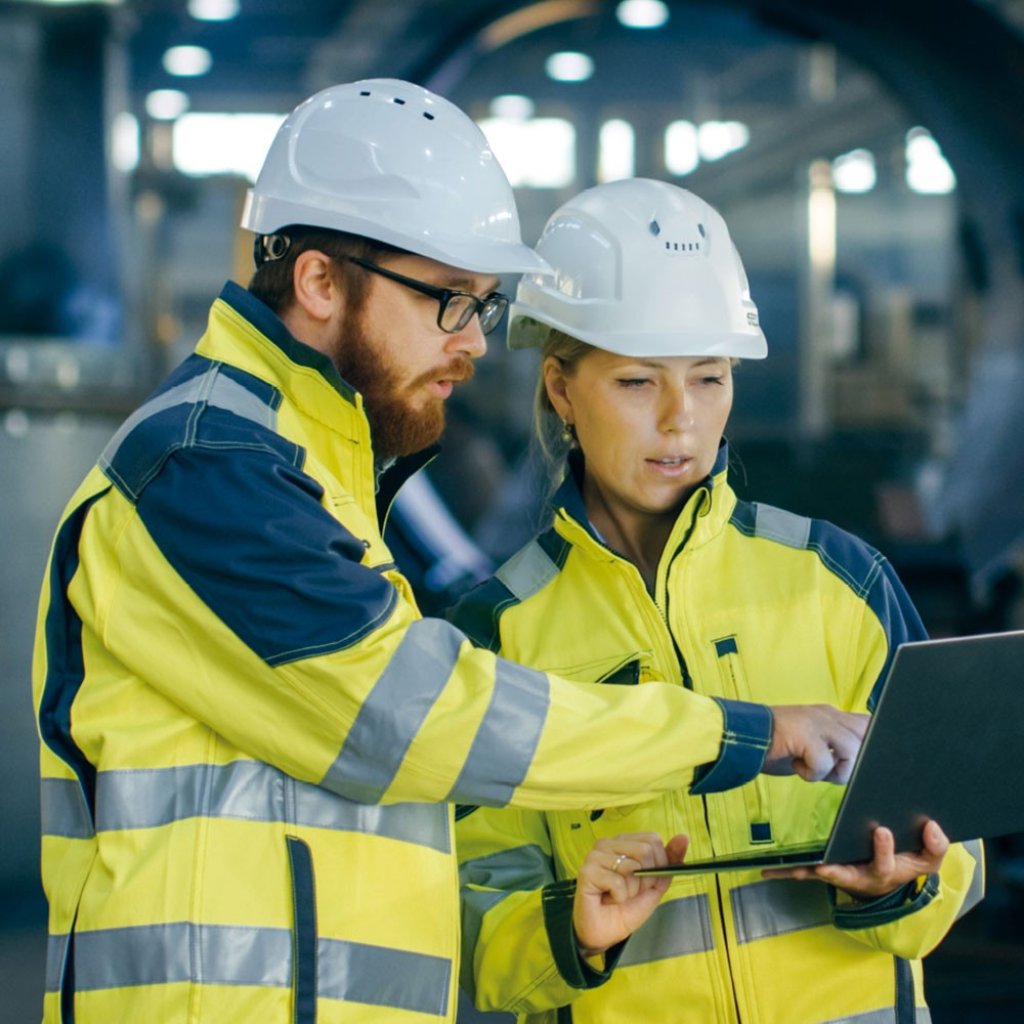Ein Mann und eine Frau in gelben Sicherheitsjacken und einem Sicherheitshelm stehen in einer Werkstatt. Die Frau hält einen Laptop und der Mann zeigt auf das Display. Lyreco unterstützt Sie mit innovativen Services im Bereich Arbeitsschutz und Sicherheit.