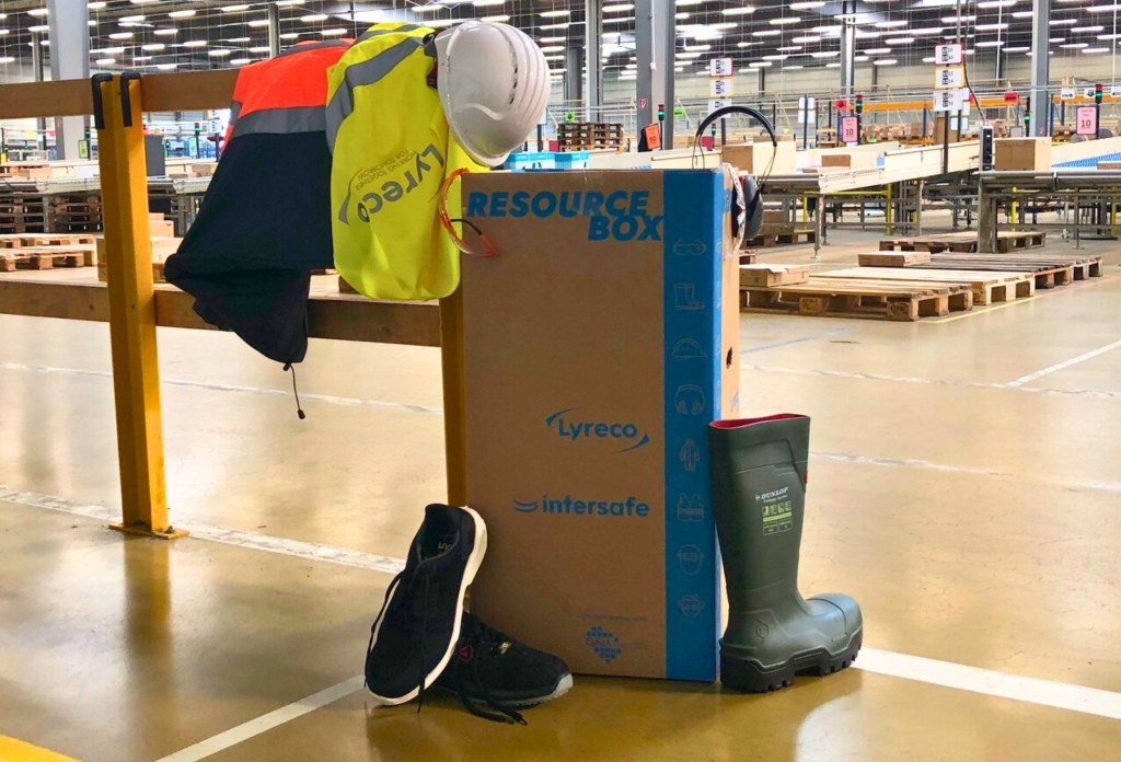 Lagerhalle von Lyreco Deutschland im Hintergrund. Im Vordergrund ist ein Geländer, auf dem sich eine Warnweste, Sicherheitsjacke und Sicherheitshelm befinden. Daneben steht eine große Box aus Karton mit der blauen Aufschrift "Resource Box". An die Box angelehnt stehen Sicherheitsschuhe in schwarz.