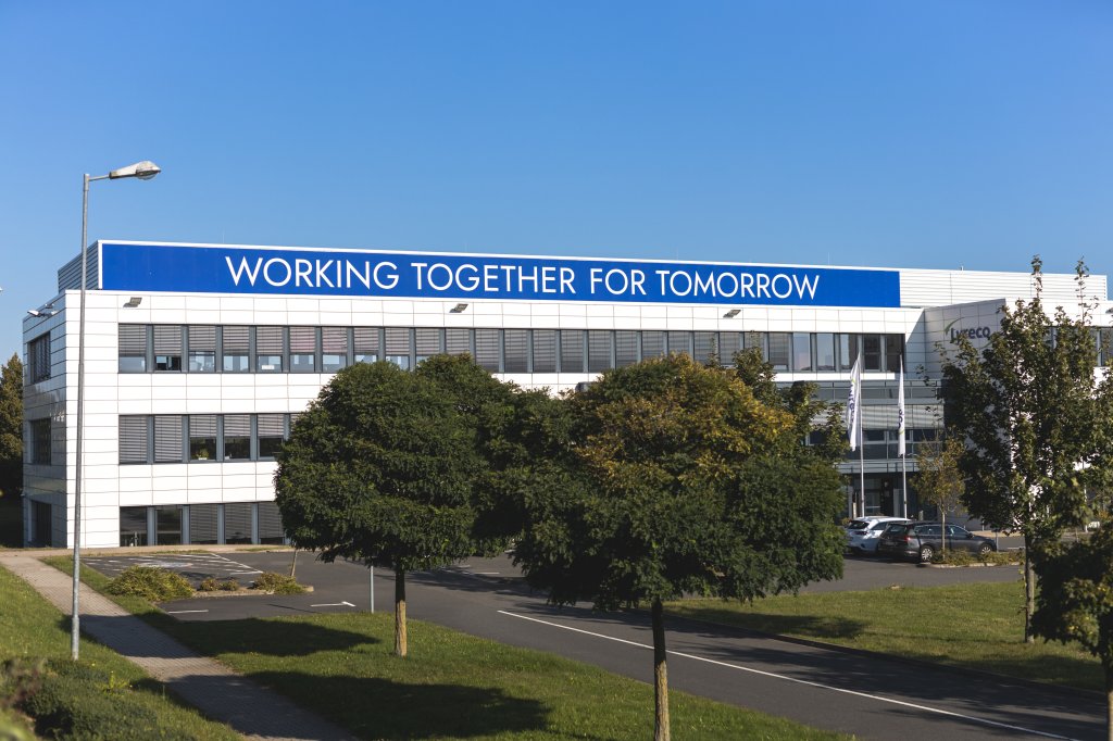 Weißes Bürogebäude von Lyreco Deutschland, oben ist in blau weißer Schrift der Satz "Working together for tomorrow" zu erkennen. im Vordergrund sind Bäume mit grünen Blättern.